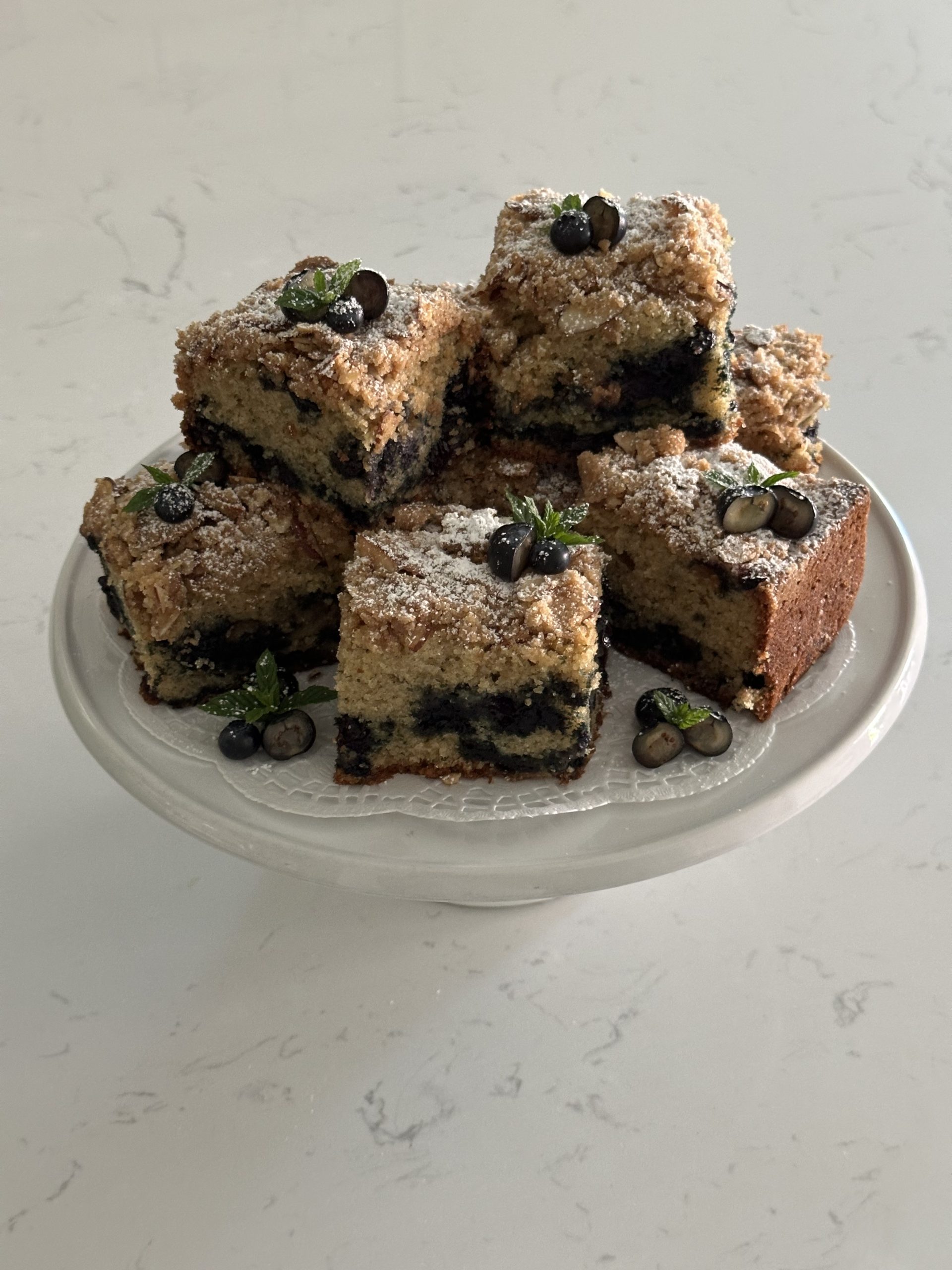 large, square pieces of blueberry mint coffee cake placed on top of a cake stand. the pieces are decorated with fresh blueberries and mint. 