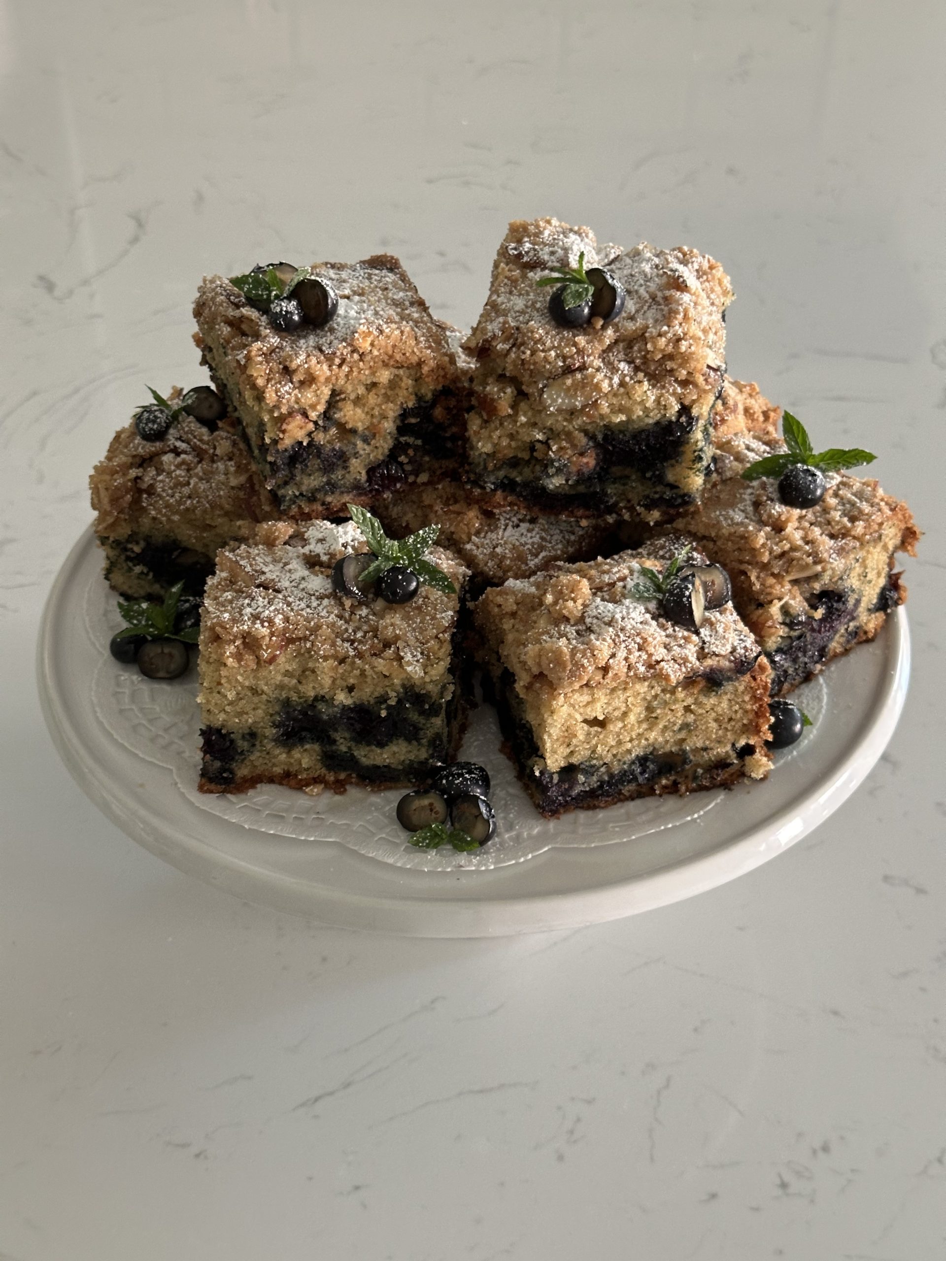 large, square pieces of blueberry mint coffee cake placed on top of a cake stand. the pieces are decorated with fresh blueberries and mint. 